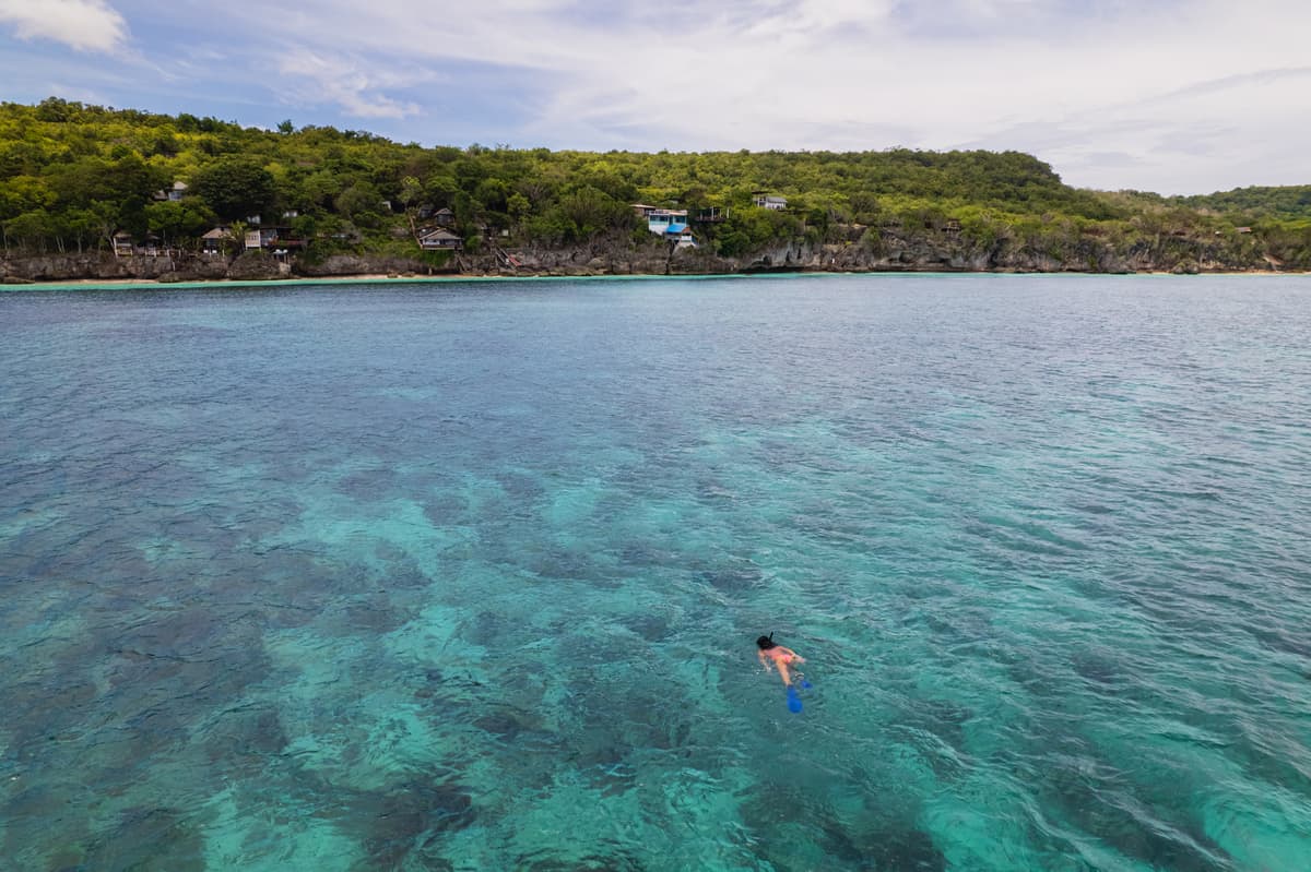 A snorkeller over the reef in clear turquoise water, the green cliff line behind